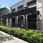 Restaurant outdoor seating under metal pergola with glass panels.