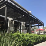 Restaurant exterior with metal awning, large windows, hedges, and umbrellas.