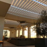 Outdoor lounge with cushioned seats, potted trees, hanging lights, louvered roof.