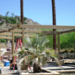 Workers building a wooden pergola frame with ladders and tools nearby.