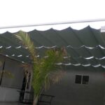 Fabric shade canopy over patio with two palm plants.
