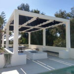 Pergola with shade panels, seating, and table beside pool.