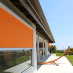 Contemporary house exterior with large window shades and hammock stand.