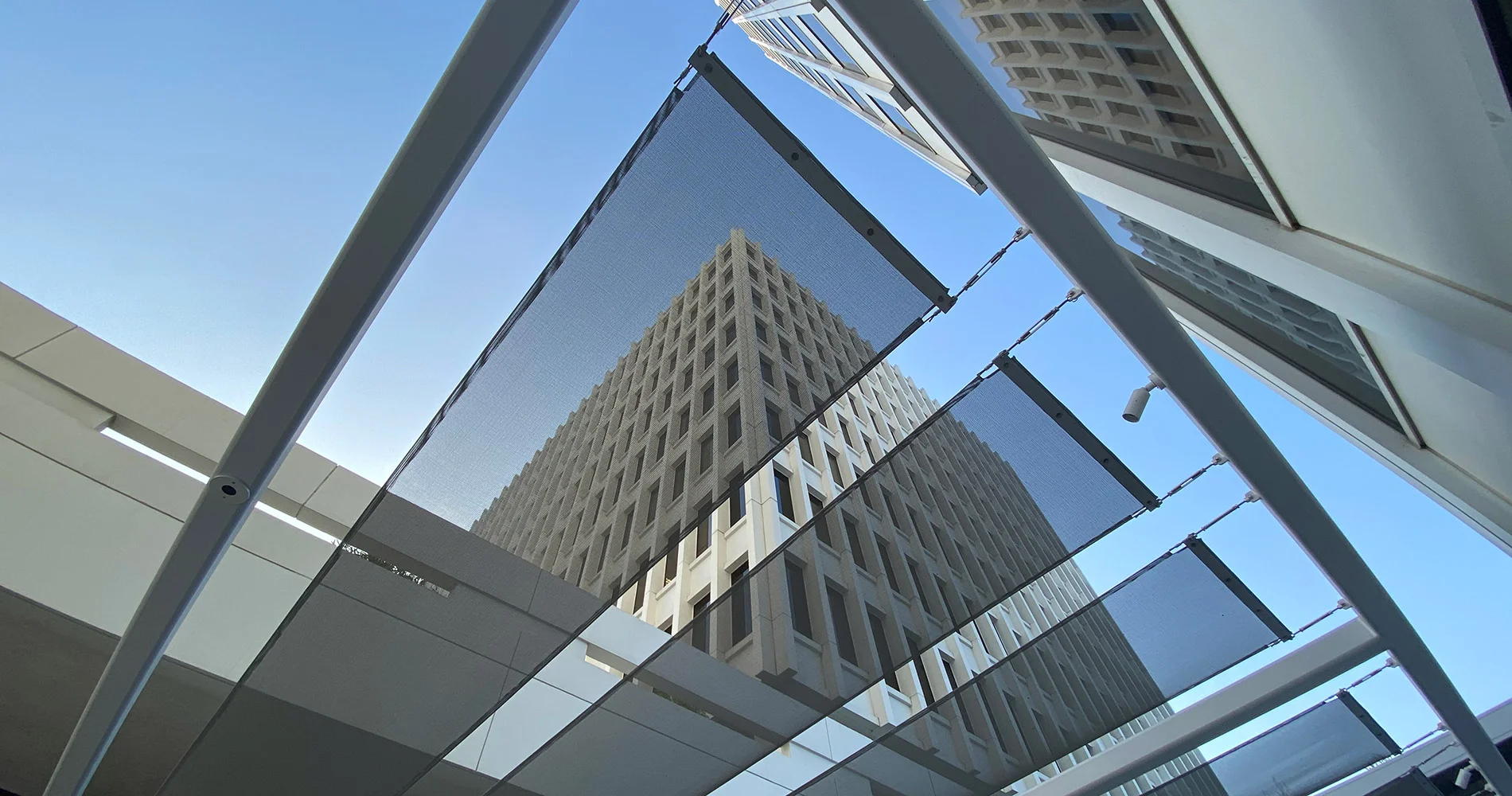View looking up at a tall, modern building seen through glass panels and a mesh canopy, with clear blue sky in the background and geometric lines framing the scene.